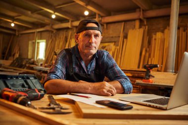 Kıdemli marangoz bilgisayarda çalışır ve atölyedeki kameraya bakar.