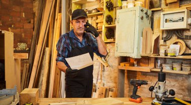 Üst düzey bir marangozun portresi mobilya yapma planlarına bakıyor ve marangozluk atölyesindeki bir müşteriyle telefonda konuşuyor.