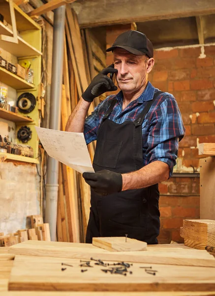 Üst düzey bir marangozun portresi mobilya yapma planlarına bakıyor ve marangozluk atölyesindeki bir müşteriyle telefonda konuşuyor.