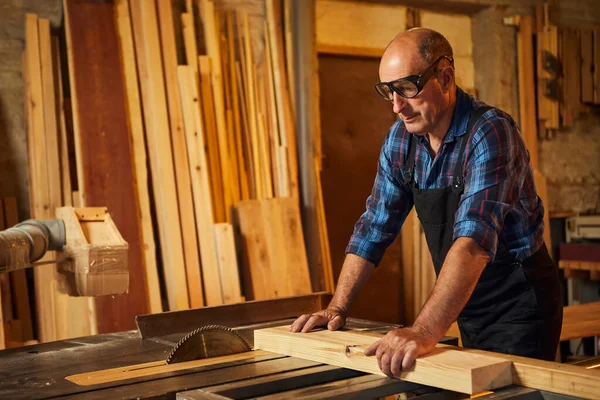 Elektrikli testere ile ahşap kesme masası. Kıdemli profesyonel marangoz üniformalı kereste fabrikasında ahşap tahta kesiyor. Testere makinesi