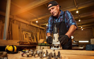 Kıdemli profesyonel marangoz üniformalı marangozluk atölyesinde elle değirmen makinesinde çalışıyor.