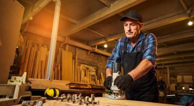 Kıdemli profesyonel marangoz üniformalı marangozluk atölyesinde elle değirmen makinesinde çalışıyor.