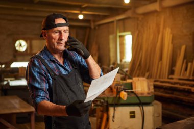 Üst düzey bir marangozun portresi planlara bakıyor ve marangozluk atölyesindeki bir müşteriyle telefonda konuşuyor.