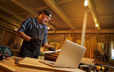 Marangoz, marangozluk atölyesinde dizüstü bilgisayarın yanında duran planlara bakıyor.