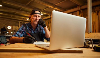 Kıdemli marangoz bilgisayarda çalışır ve atölyedeki müşteriyle telefonda konuşur.