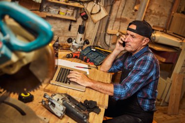 Kıdemli marangoz bilgisayarda çalışır ve atölyedeki müşteriyle telefonda konuşur.