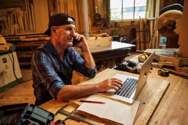 Kıdemli marangoz bilgisayarda çalışır ve atölyedeki müşteriyle telefonda konuşur.