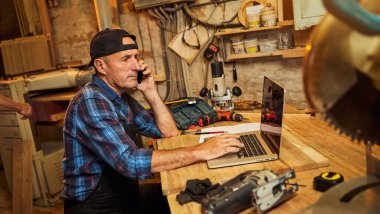 Kıdemli marangoz bilgisayarda çalışır ve atölyedeki müşteriyle telefonda konuşur.