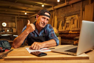 Gülümseyen marangoz portresi bilgisayarda çalışır ve atölyede parmağını kaldırır.