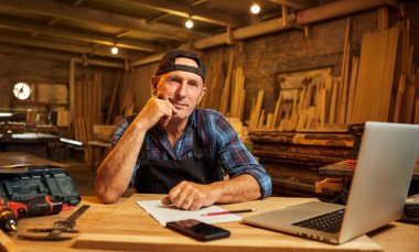 Kıdemli marangoz bilgisayarda çalışır ve atölyedeki kameraya bakar.