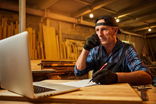 Senior carpenter works on the computer and talks phone with client in the workshop