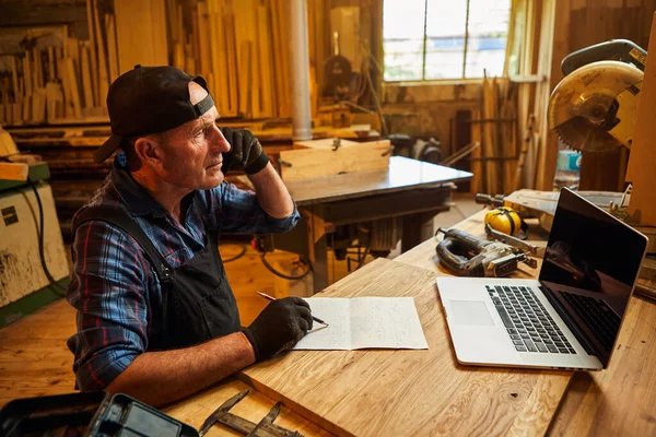 Senior carpenter works on the computer and talks phone with client in the workshop