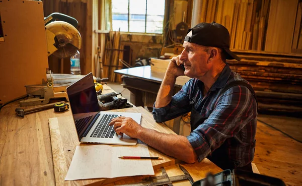 Kıdemli marangoz bilgisayarda çalışır ve atölyedeki müşteriyle telefonda konuşur.