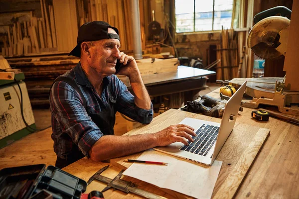 Kıdemli marangoz bilgisayarda çalışır ve atölyedeki müşteriyle telefonda konuşur.