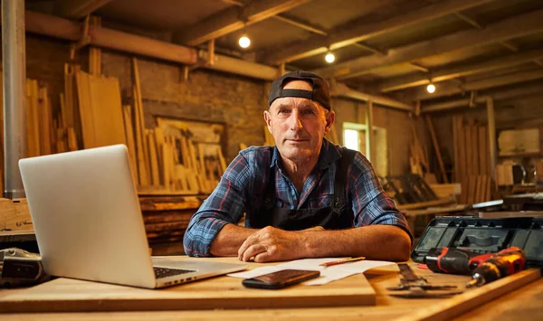 Senior carpenter works on the computer and look at the camera in the workshop