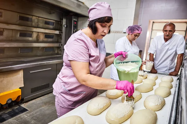 Meslektaşlarının arka planındaki fırında pişirmek için üniformalı profesyonel fırıncı tereyağlı ekmek hamuru sürüyor.