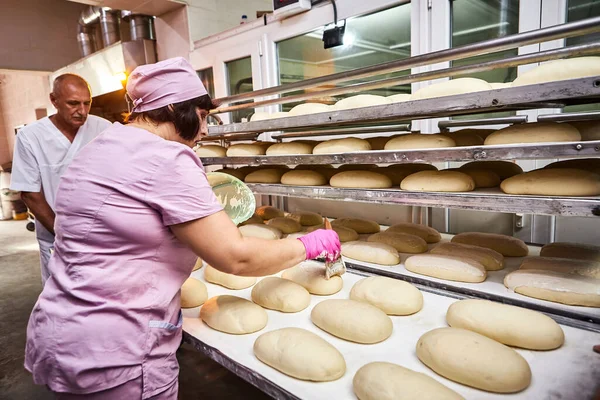 Meslektaşlarının arka planındaki fırında pişirmek için üniformalı profesyonel fırıncı tereyağlı ekmek hamuru sürüyor.