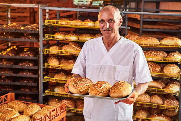 stock image Baker holds a tray with fresh hot bread in his hands against background of shelves with pastries in bakery. Industrial production of bread