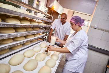 Meslektaşlarının arka planındaki fırında pişirmek için üniformalı profesyonel fırıncı tereyağlı ekmek hamuru sürüyor.