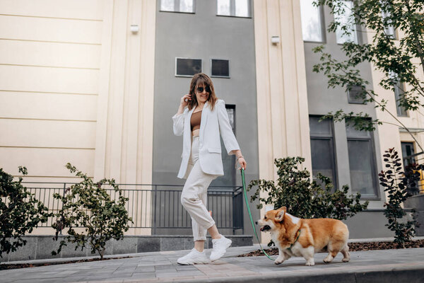 Beautiful Woman wearing white suit and sunglasses walking down the street with Welsh Corgi Pembroke dog