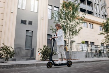 Beautiful young woman in sunglasses and white suit is riding on her electric scooter near modern building in city and looking away