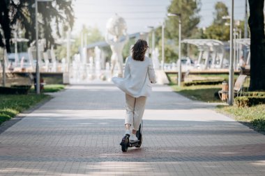 Beautiful young woman in white suit is riding to work on her electric scooter in city parkland