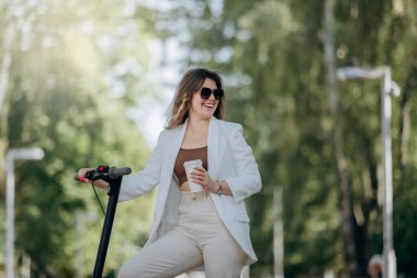 Beautiful young woman in white suit and sunglasses standing with her electric scooter in city parkland and drinking coffee