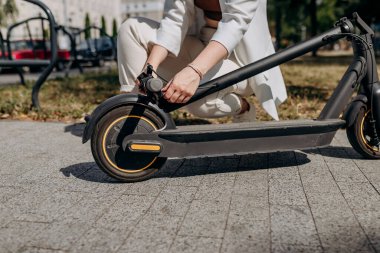 Close up of woman in white suit folding her electro scooter after ride while standing in city parkland