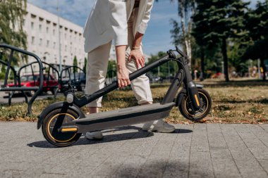 Close up of woman in white suit folding her electro scooter after ride while standing in city parkland