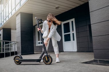 Pretty woman in white suit folding her electro scooter after ride while standing on background of modern building