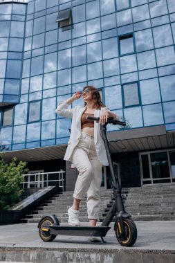 Beautiful young woman in sunglasses and white suit standing with her electric scooter near modern building and looking away 