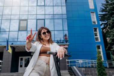 Beautiful young woman in sunglasses and white suit standing with her electric scooter near modern building and looking at camera and showing sign V