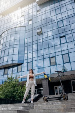 Beautiful young woman in sunglasses and white suit standing with her electric scooter near modern building and looking away 