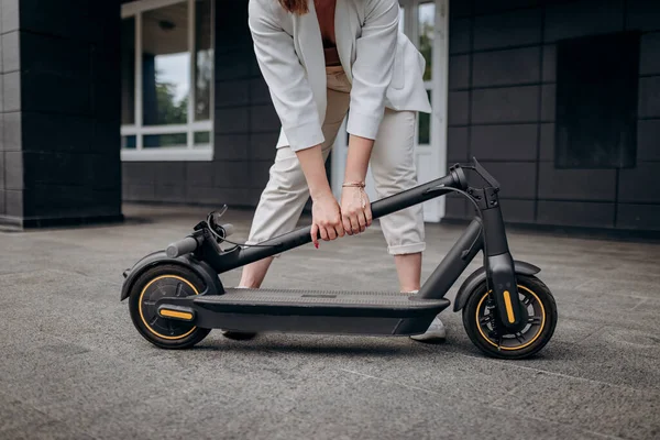 Close up of woman in white suit folding her electro scooter after ride while standing on background of modern building