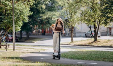Happy smiling woman traveler is riding her electro scooter in city parkland with dog Welsh Corgi Pembroke in a special backpack