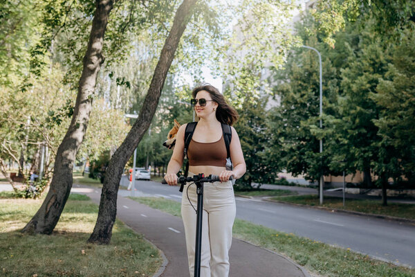 Счастливая улыбающаяся женщина путешественник едет на электроскутере в городском парке с собакой Уэльс Корги Пемброк в специальном рюкзаке