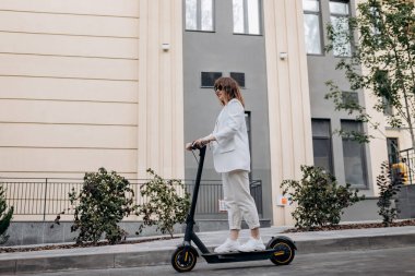 Beautiful young woman in sunglasses and white suit is riding on her electric scooter near modern building in city and looking away