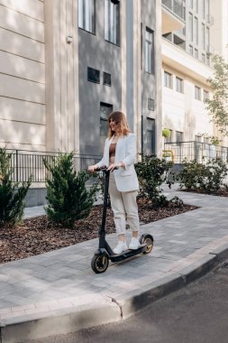 Beautiful young woman in sunglasses and white suit is riding on her electric scooter near modern building in city and looking away
