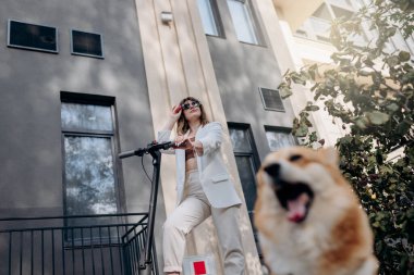 Beautiful young woman in white suit standing with her electric scooter and COrgi Dog near modern building in city and looking away