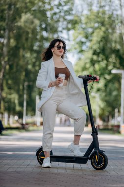 Beautiful young woman in white suit and sunglasses standing with her electric scooter in city parkland and drinking coffee