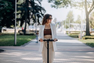 Beautiful young woman in white suit is riding to work on her electric scooter in city parkland 