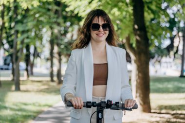 Beautiful young woman in white suit is riding to work on her electric scooter in city parkland 