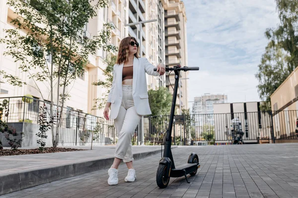 Beautiful young woman in sunglasses and white suit standing with her electric scooter near modern building in city and looking away