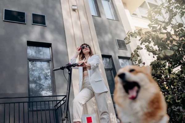 Beautiful young woman in white suit standing with her electric scooter and COrgi Dog near modern building in city and looking away