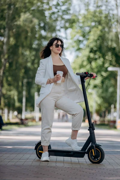 Beautiful young woman in white suit and sunglasses standing with her electric scooter in city parkland and drinking coffee