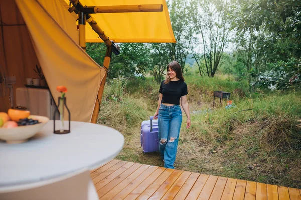 Happy woman traveler with luggage moving towards bedroom of cozy glamping house. Luxury camping tent for outdoor summer holiday and vacation. Lifestyle concept