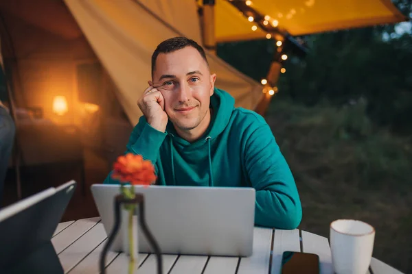 Happy man freelancer using a laptop on a cozy glamping tent in a summer evening. Luxury camping tent for outdoor holiday and vacation. Lifestyle concept