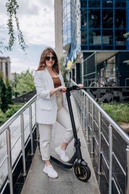 Beautiful young woman in sunglasses and white suit standing with her electric scooter near modern building and looking at camera