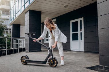 Pretty woman in white suit folding her electro scooter after ride while standing on background of modern building