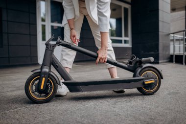 Close up of woman in white suit folding her electro scooter after ride while standing on background of modern building